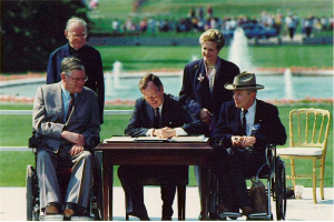 President Bush is seated at a table outside, signing a document. Reverend Harold Wilke and Sandra Parrino stand behind him, Evan Kemp and Justin Dart are to the left and right of him