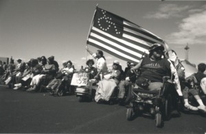 Protestors - many using wheelchairs and one man with an american flag that has the stars in the shape of a wheelchair