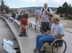 photo of a gentlemen in a wheelchair reading a sign at Yellowstone National Park - there are a dozen or so other people behind him some on benches and some standing