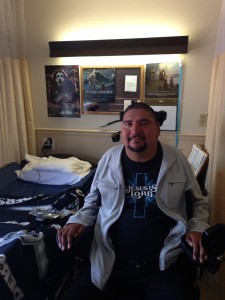 Juan Jose sits in an electric wheelchair in his room. You can see his bed and some posters up behind him.