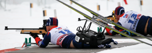 2 Biathlon atheltes with their guns taking shots laying down in the snow wearing their skis