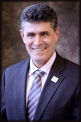 head shot of Joe wearing a suit and tie and pin, smiling