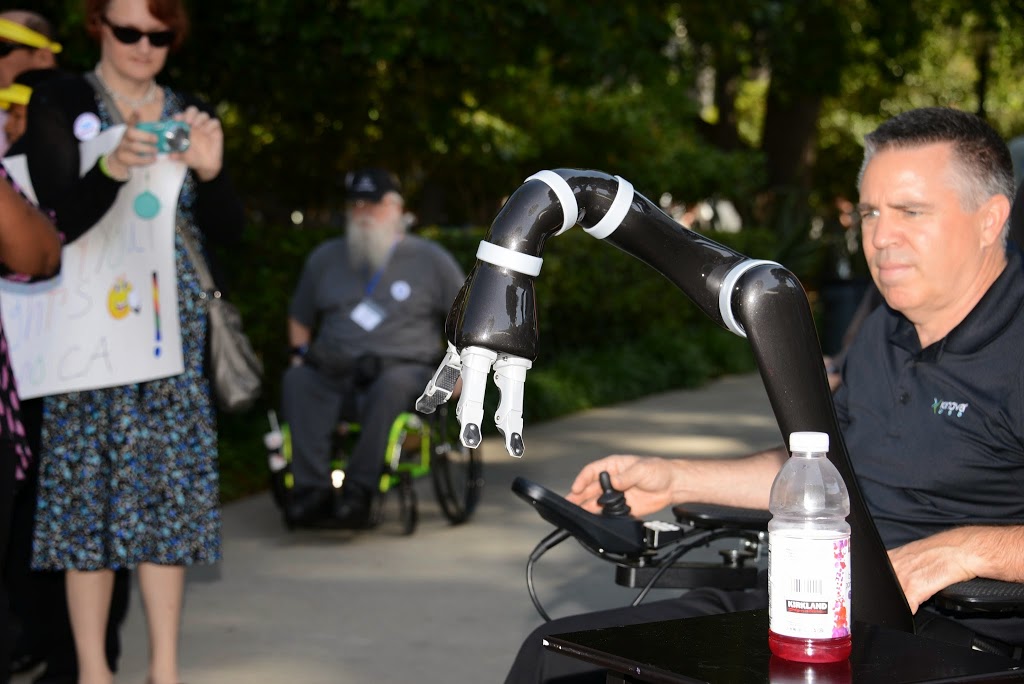 a man using a power wheelchair uses the JACO robotic arm device to reach for a bottled beverage.