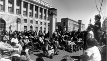 Black and white photo of a bunch of people including many that use wheelchairs listening to a woman speak outside government building