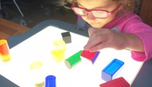 Olivia using her light box wearing pink glasses and looking at different colors and shpaes of blocks on the box