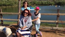 Picture of Aleta with her two daughters she is sitting in a wheelchair smiling and her two daughters are behind her standing and there is a lake behind them