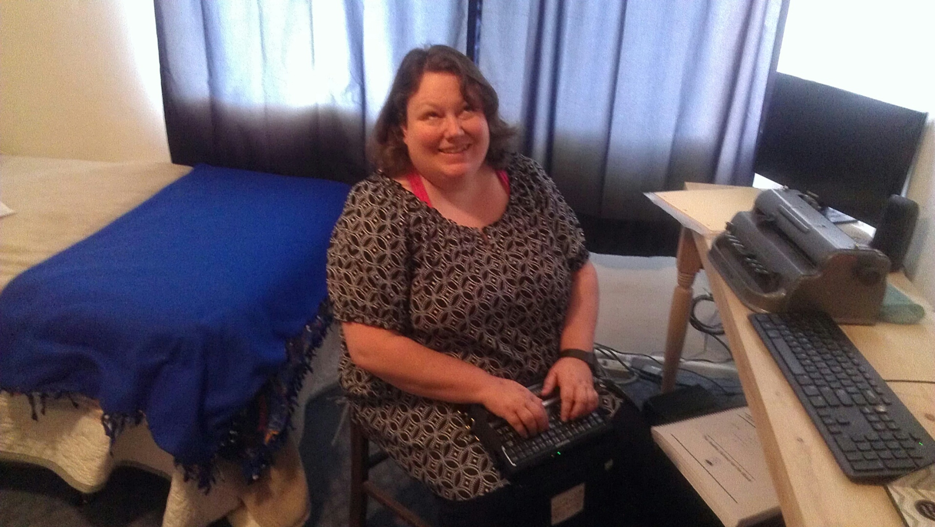 Picture of Nicole seated and smiling at her computer desk and using a Braille note. 