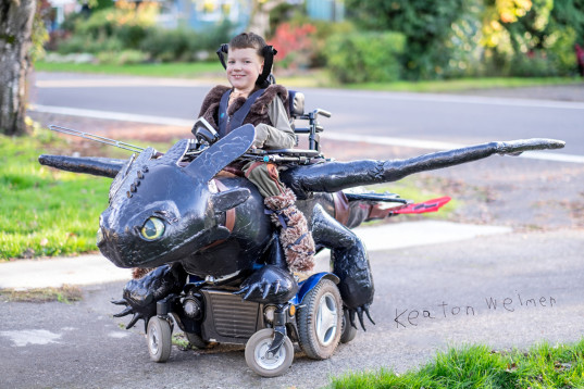 picture of young boy named Keaton Welmor and his wheelchair is covered in an elaborate flying froglike creature costume and he is smiling 