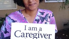 picture of a smiling woman seated and holding a placard that says I am a caregiver for my mom