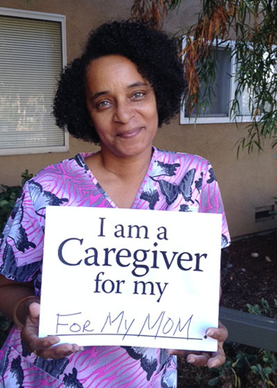 picture of a smiling woman seated and holding a placard that says I am a caregiver for my  mom