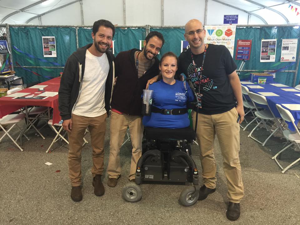 kim lathrop and her TOM cohort. She is wearing a blue shirt and uses a wheelchair and three other gentleman are smiling and standing with their arms around her.