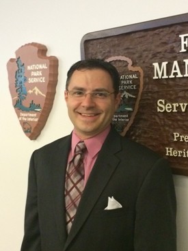 picture of man smiling in front of park service plaque wearing a suit and tie