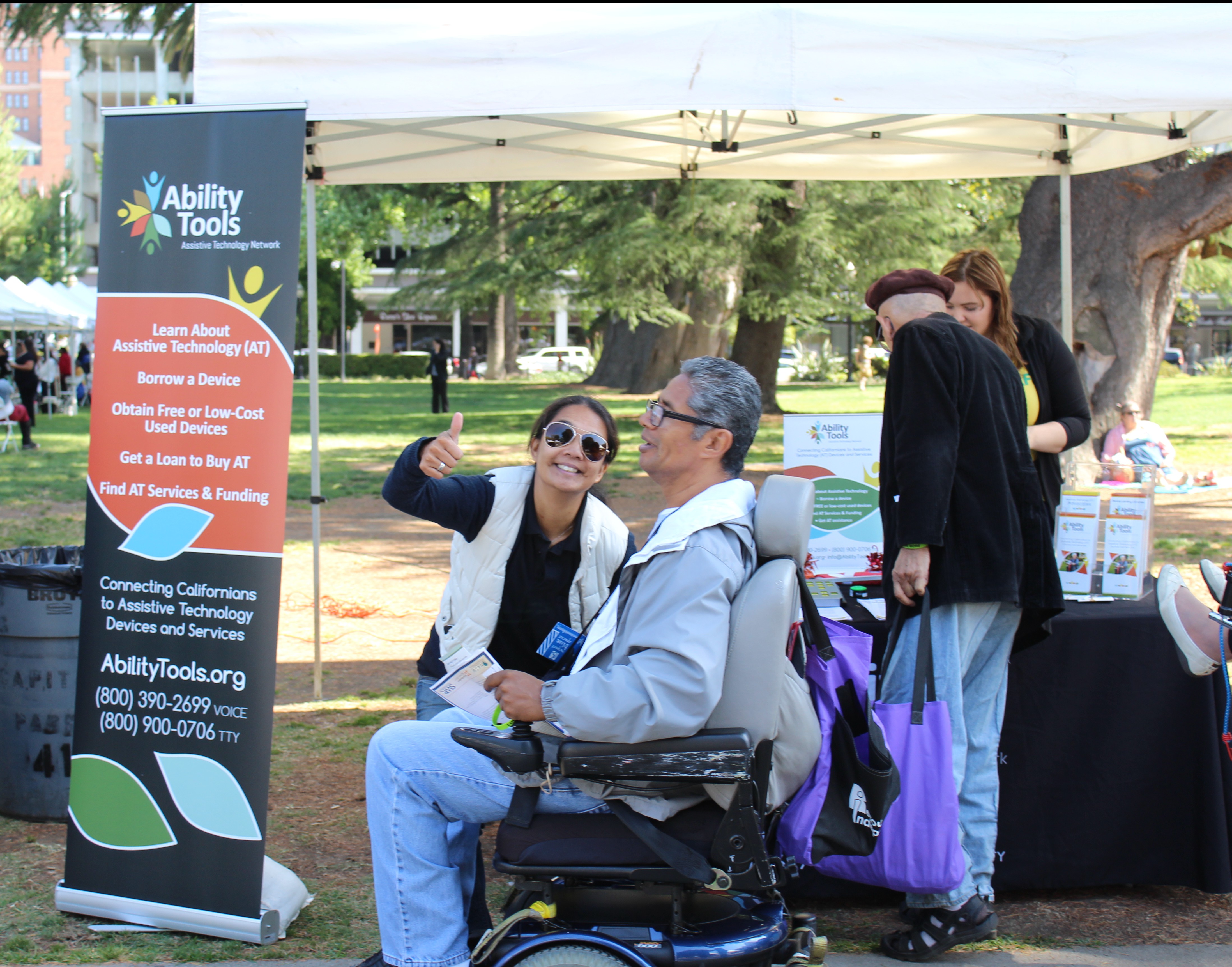 Women with sunglasses giving the photographer a thumbs up while she speaks with a man in a power chair in front of the Ability Tools booth 