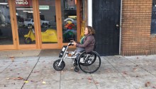 Women in wheelchair with firefly attached to the front zooming past a store front