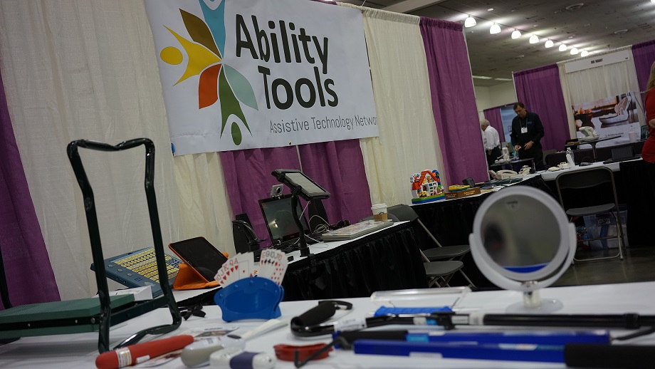 A photo of the AT booth. There is an Ability tools banner hanging with purple and white curtains. On the table there are various AT devices for people to try. 