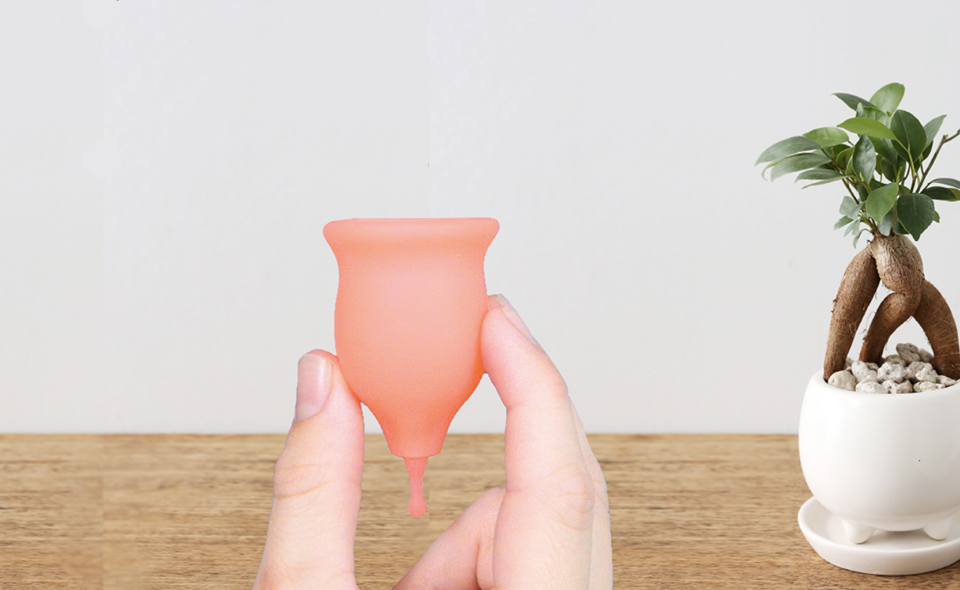 There is a gray wall in the background with a wooden table and small bonsai plant. A hand is holding up a keela cup. The cup is pink and shaped like a small inverted bell with a long stem at the bottom.