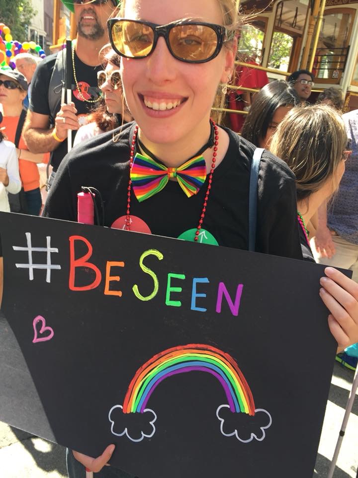 a Caucasian woman wearing sunglasses and a rainbow bowtie holding a pink cane and a sign that says be seen with a rainbow.