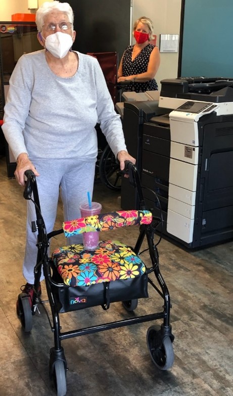 An older woman wearing glasses and a facemask utilizes a floral designed walker in an office building.