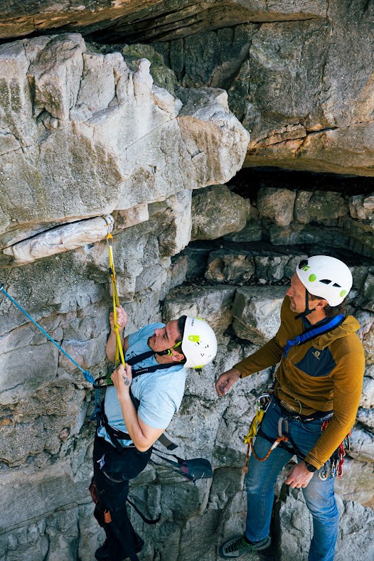 Two climbers wearing gear, are traversing a cliff side. One is actively climbing and the other stands nearby looking at the cliffside.