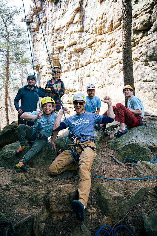 A group of people wearing climbing gear are perched on a rocky hillside with a cliff in the background. They are all smiling and posing at the camera.