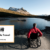 A person wearing a red jacket, sits in a wheelchair beside a lake looking at a sunny snow capped mountain view. Beside the Where it's AT logo, text reads Accessible Travel - AbilityTools.org