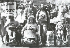 a black and white picture shows a crowd of people, some of whom use wheelchairs, one man has a service dog with him. They are children and adults and appear to be chanting or singing. One person holds a sign that says, “We shall overcome.”