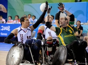 Picture of 3 men in wheelchairs fighting for control of the rugby ball all with their hands up in the air. Shirts say US and Australia. 