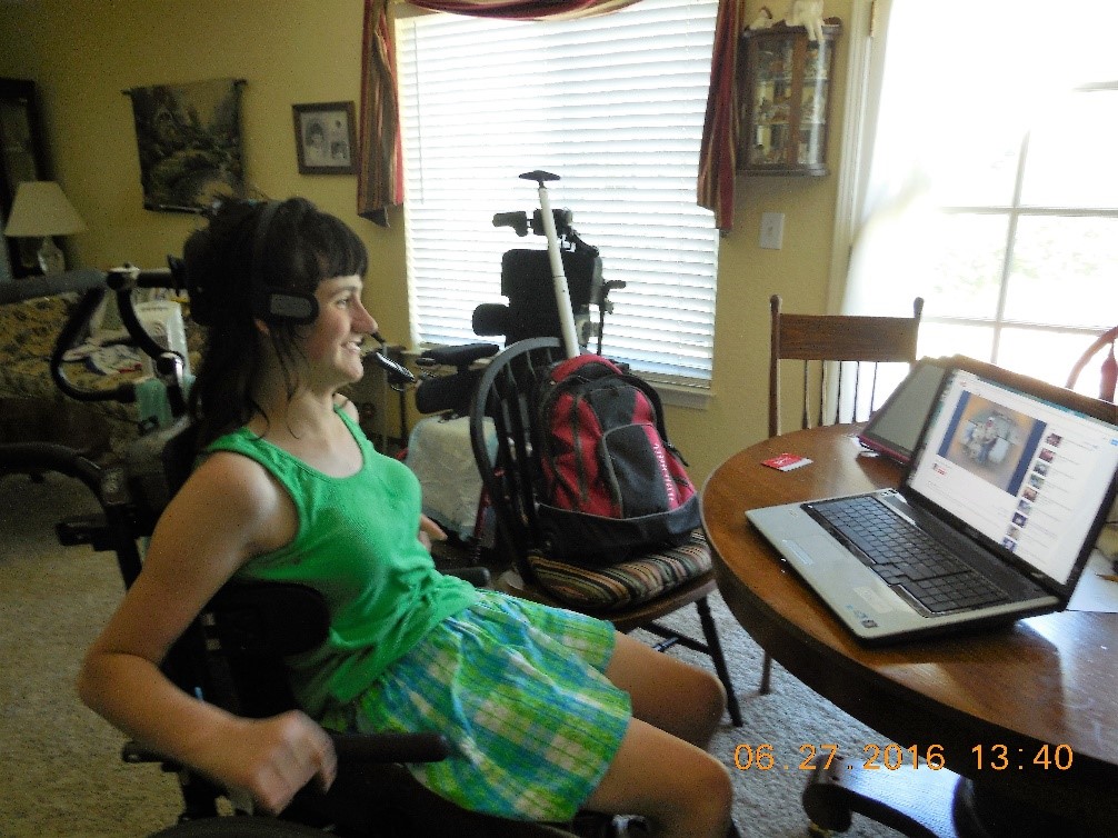 catherine stiting at a dining room table in her wheelchair and in front of computer using the mouse heaadset