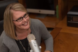 Picture of blonde women in glasses smiling while speaking into a microphone 