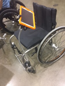Manual chair with long arm attached to a mounting system holding an Ipad with orange case.