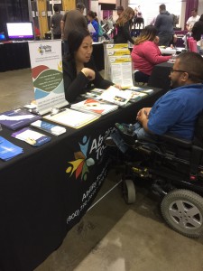 Women sitting behind a table speaking with a young man in a power chair 