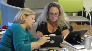 Two women sitting with an ipad,  one women is showing the other how to access a speech app.