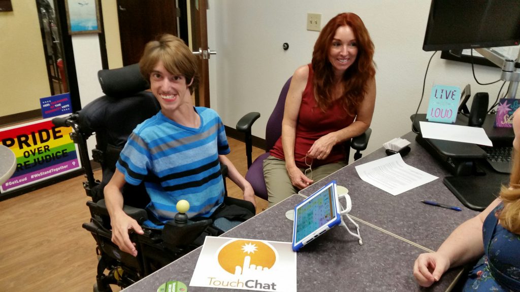 A young man using a power chair is sitting next to his mom in an office. There is an iPad on the table with a speech app.