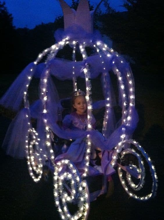 Little girl dressed in a beautiful pink princess dress sits in her power chair decorated to look like a big carriage equipped with twinkling lights and Tull 
