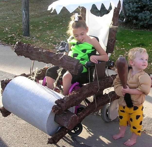 little girl dressed in green cave women outfit with a bone in her ponytail. Her power chair is decorated to look like a prehistoric car like the ones featured in the Flintstones