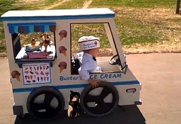 Little boy wears a white hat that reads "busters ice cream" along with a bow tie. The little boy pushes his manual chair that is designed to look like an ice cream truck stocked with faux ice cream cones in the back window