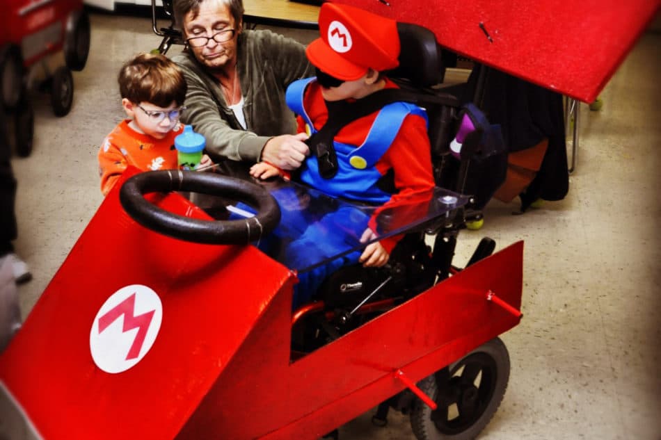 Little boy dressed in blue overalls and a red hat and mustache dressed to look like Mario, sits in his power chair decorated to look like a Red go Kart