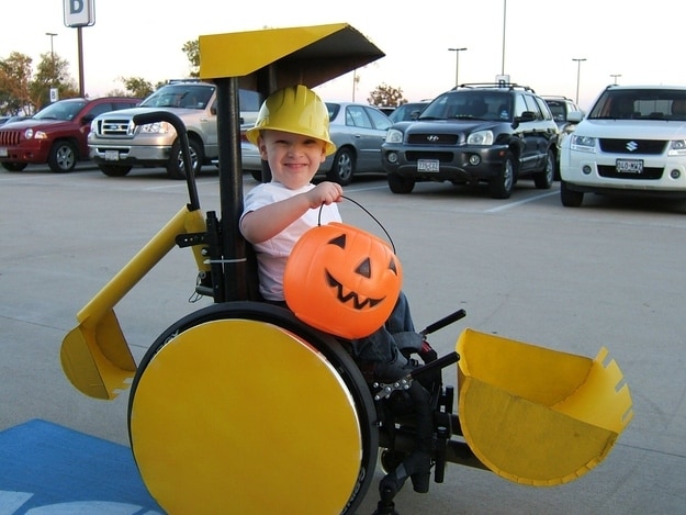 Little boy wearing a hard hat and holding up a jack-o-lantern pale while he sits in his power chair that is decorated to look like a little yellow bulldozer 