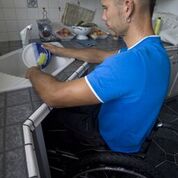 A gentleman using a wheelchair is pulled up at his roll under sink washing a plate. 