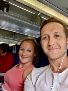 A woman wearing a pink top and a man wearing earbud headphones are sitting together on an airplane.