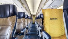 The photo is of the inside of an empty airplane. One of the seats has a yellow ADAPTS sling covering it.