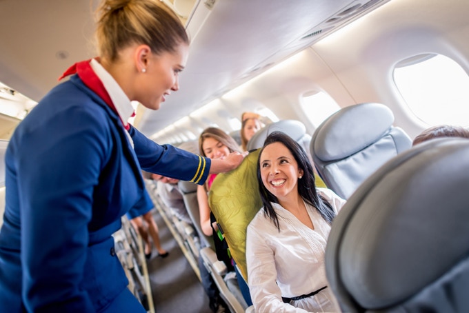 A woman is sitting on an airplane seat with the ADAPTS sling beneath her talking to a flight attendant. 