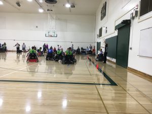 Two Rugby Wheelchair teams, one wearing blue the other green, are playing on a basketball court.