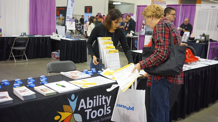 A woman with dark brown hair is standing behind the Ability Tools booth. She is talking with a woman handing her materials. 