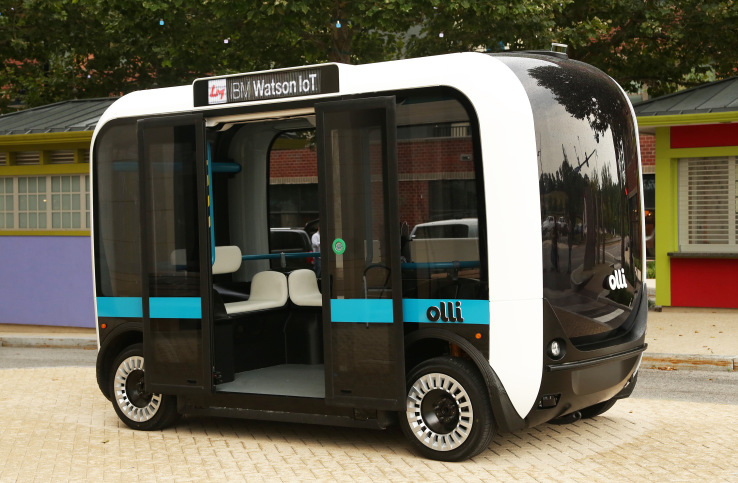 the black,white, and blue Olli bus sits in city scene with boors open welcoming passengers 