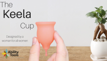 A gray wall with a wooden table and small bonsai plant. A hand is holding up a pink Keela Cup. It is shaped like an inverted bell with a long pullstring on the bottom. The text reads 'The Keela Cup Designed by a woman for all women". Th Ability Tools logo is on the bottom left.