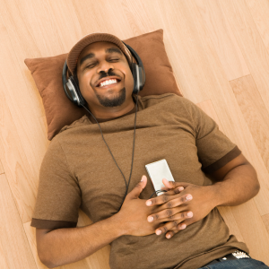 Man of color laying on wood floor with brown pillow, smiling with eyes closed, wearing brown hat and shirt. Wearing headphones and holding a player.