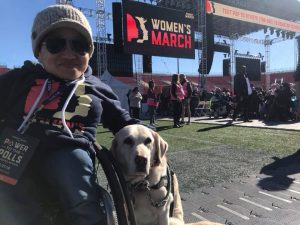 Photo of Mia, woman of Asian heritage wearing sunglasses and a beanie, seated in her wheelchair with service dog Ari by her side at the Women's March in Washington.