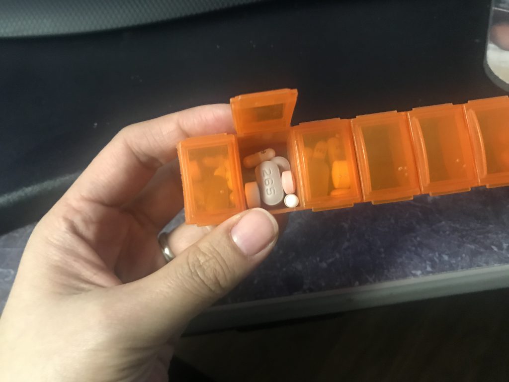 Photo of Kyla’s left hand holding a pill holder with 7 small boxes. One of the box lids is lifted to show pills inside.