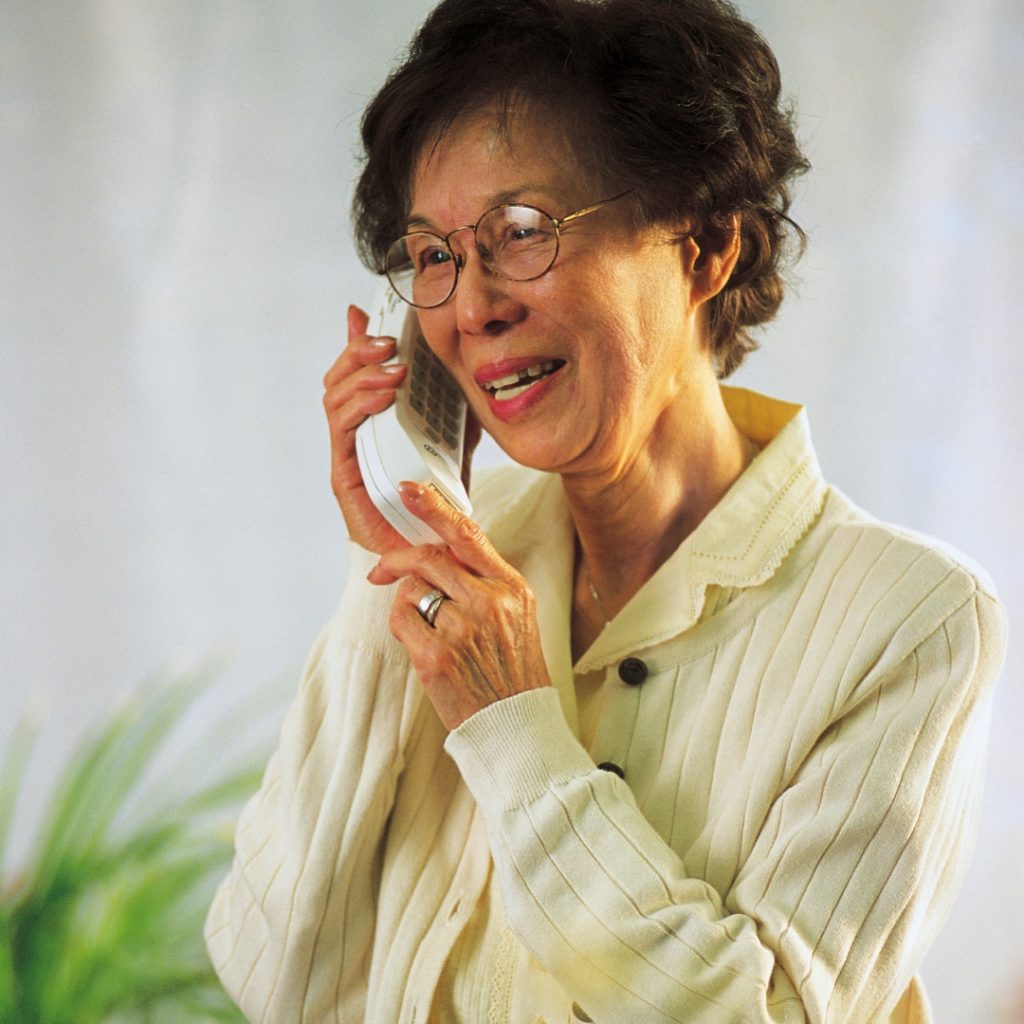 Indoor photo of an elder Asian woman on a landline telephone wearing eyeglasses and a beige cardigan. smiling.